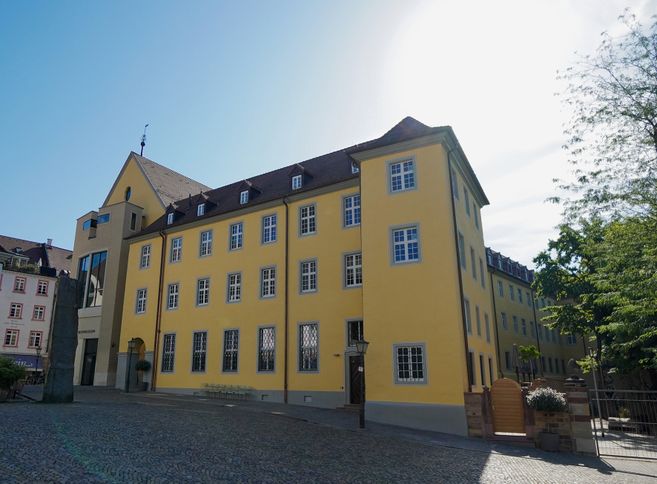 Außenansicht des Augustinermuseums in Freiburg mit gelber Fassade und blauen Fenstern unter klarem Himmel.