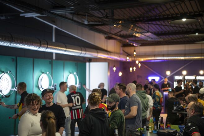 Dartzentrum Freiburg, Dartboards mit Beleuchtung an den Wänden, Bar und Lounge mit warmer Beleuchtung, Menschen, die Darts werfen, zuschauen oder miteinander sprechen. 