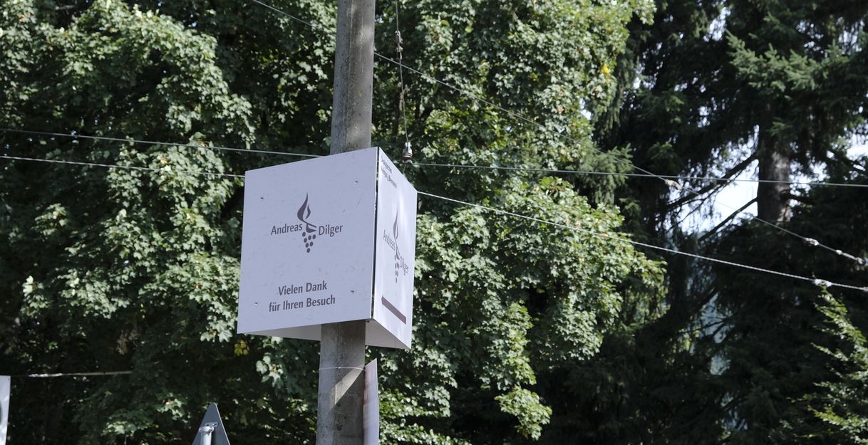 Ein Schild mit der Aufschrift 'Andreas Dilger' und 'Vielen Dank für Ihren Besuch' an einem Pfosten vor Bäumen.